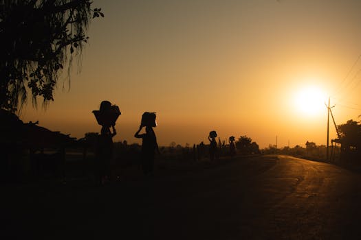 Dramatic sunset scene with silhouetted figures carrying baskets along an Indian road.